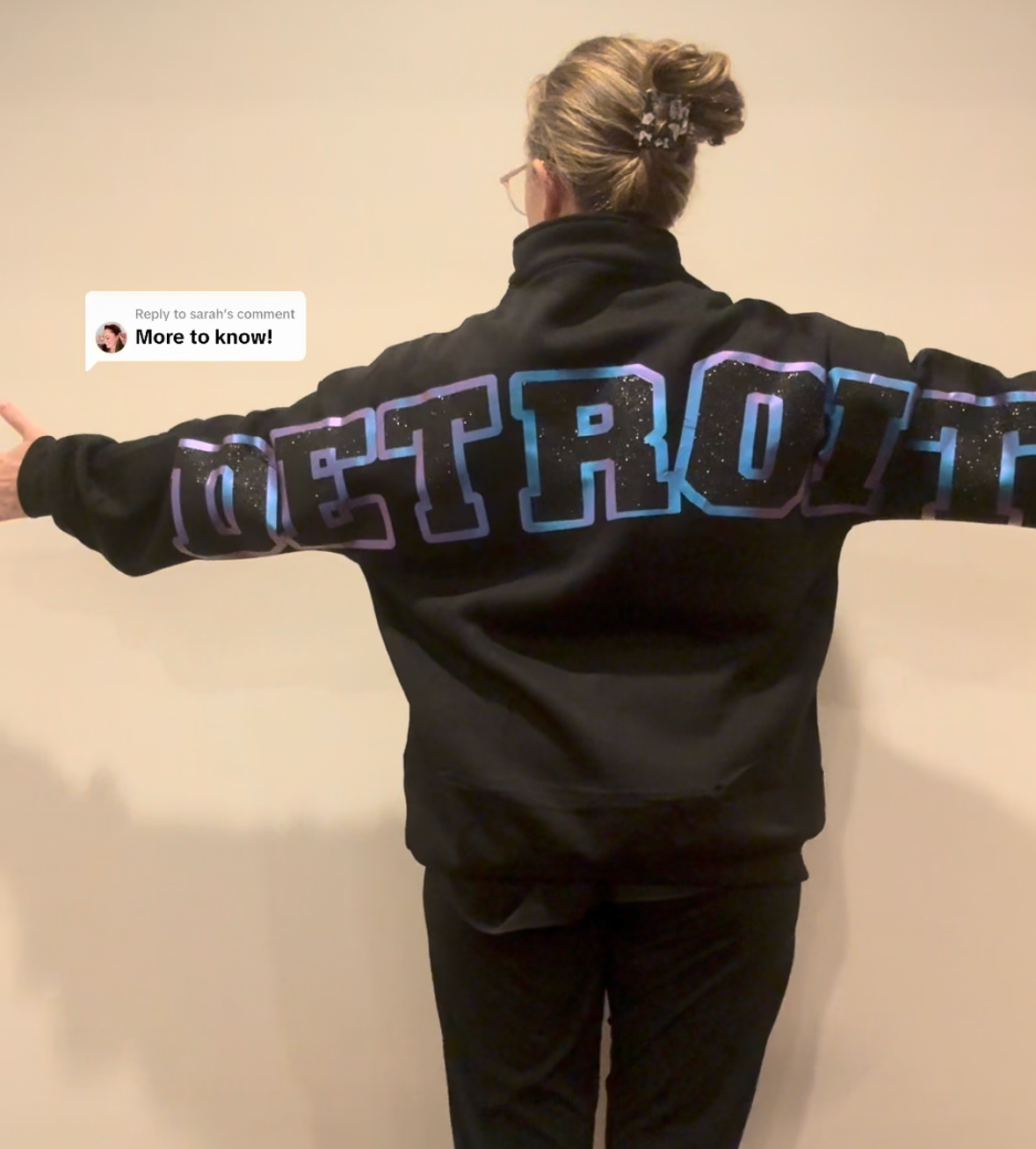 A person shown from behind wearing a black Detroit Tigers sweatshirt with large blue and white lettering across the back, standing indoors with arms outstretched. The photo appears to be a screenshot from a video call or social media-style frame, with phone status icons, a timer, and a video player bar visible.